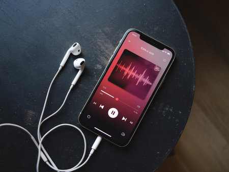 A smartphone with earphones resting on a dark table, displaying a music app interface.の素材