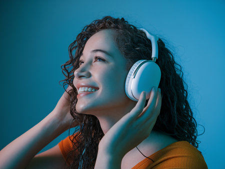 Smiling young woman with curly hair enjoys music with white headphones against a blue background.の素材