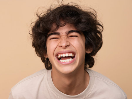 A joyful young boy with curly hair laughing heartily against a soft beige background.の素材