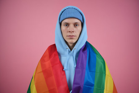 Young Caucasian male wrapped in a rainbow flag, wearing a blue hoodie and beanie against a pink background.の素材