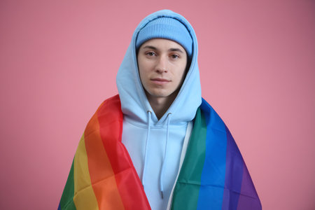 A young Caucasian man wraps himself in a rainbow flag, standing against a pink backdrop.の素材
