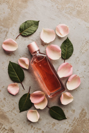 A pink glass perfume bottle surrounded by rose petals and green leaves on a textured surface.の素材