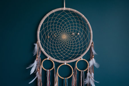 A beautifully crafted dream catcher with delicate feathers and beads hanging against a teal wall.の素材