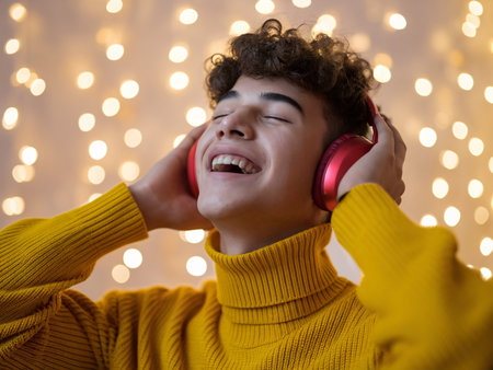 Young male with curly hair enjoying music with headphones, wearing a cozy yellow sweater amidst festive lights.の素材