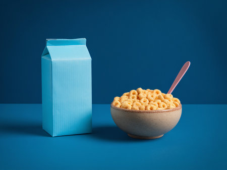 A bowl of yellow cereal with a pink spoon next to a blue milk carton on a vibrant blue background.の素材