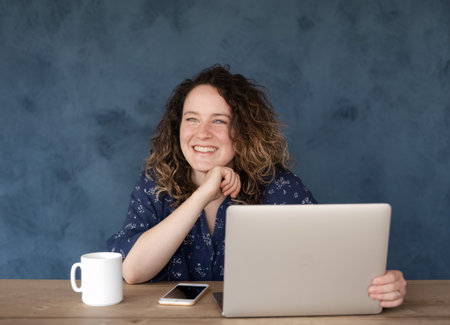 Smiling woman with curly hair working on a laptop, enjoying her coffee in a cozy workspace.の素材