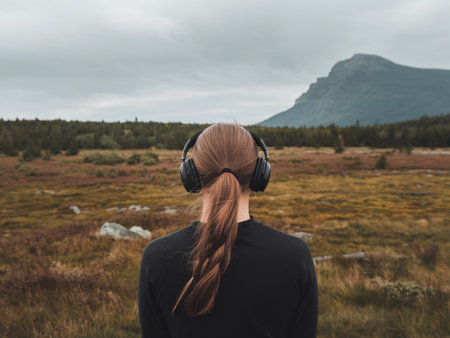 Young woman with long hair and headphones enjoying nature view in a serene landscape.の素材