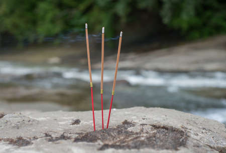 Three steaming buddhist incense sticks in the stone on the river bank. Selective focus.の写真素材