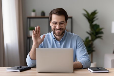 Happy male freelancer waving at camera, greeting client, making video call, chatting with colleagues, sitting at desk in office.の素材