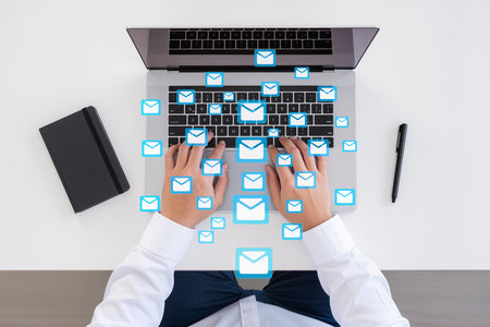 Businessman sitting at desk using laptop against email icons on white backgroundの素材
