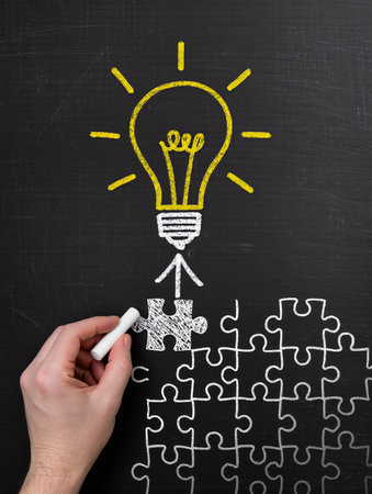 Hand drawing a light bulb on a blackboard with a piece of puzzleの素材