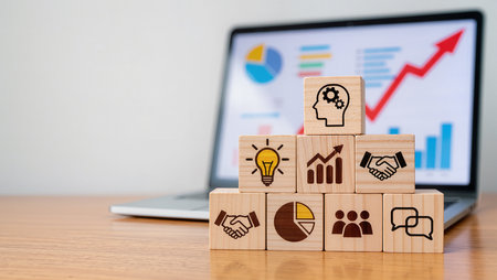 Wooden blocks with business icons on a desk in front of a laptopの素材