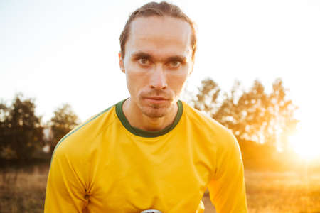 Attractive serious young European man, standing in a meadow, wearing yellow sportswear looking at camera on sunset flareの写真素材