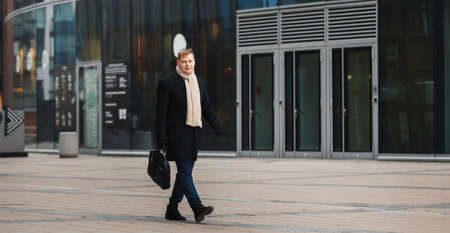 Side view of elegant man in black coat walking on sidewalk against contemporary building.の写真素材