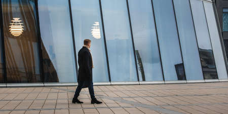 Side view of elegant man in black coat walking on sidewalk against contemporary building. の写真素材