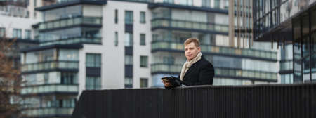 Elegant confident businessman in coat holding gadget and standing on terrace with urban background.の写真素材