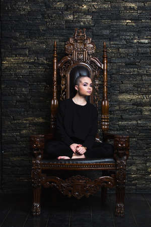 Beautiful young model with makeup, wearing black sweatshirt, sitting in a big chair throne looking expressively at cameraの写真素材