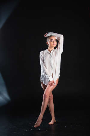 Beautiful alluring model with shaved head standing and posing nude in white shirt and glasses in studio on black backgroundの写真素材