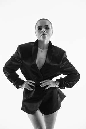 Young nude woman with shaved head covering body with short shirt looking at camera on white.