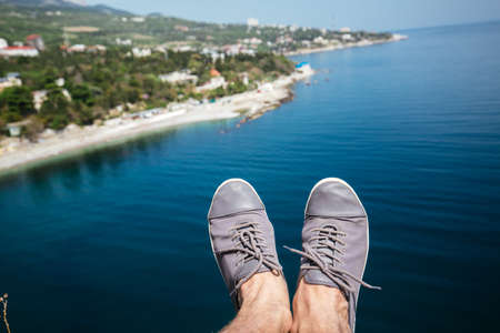 Male legs in grey sneakers lit by sun over cliff against backdrop of sea and beach.