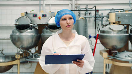 Controller checking confection factory. Young female in uniform making notes in folder while inspecting laboratory in confection factory.の写真素材