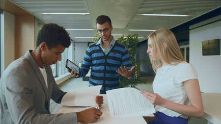 Side view of black man with coworkers at table reading papers and using tablet in team at table.の写真素材