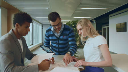 Side view of black man with coworkers at table reading papers and using tablet in team at table.の写真素材