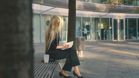 Side view of elegant businesswoman sitting on bench in patio and writing in notepad while working.の写真素材