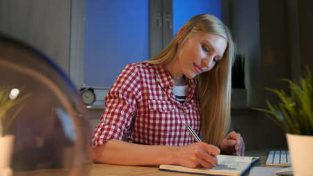 Lively young blond woman in casual checkered shirt, sitting at wooden desk with notebook holding metal pen and looking at camera smiling at night in dark room.の写真素材