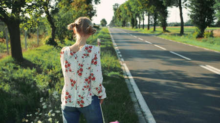 Young woman on countryside road. Back view of young female with red suitcase standing on roadside while travelling through countryside.の写真素材