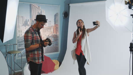 Pretty black model making selfie on smartphone during photo shoot in modern studio. The photographer asking her to start working.の写真素材
