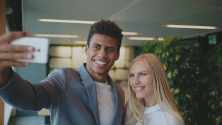 Cheerful black man with laughing blond woman taking selfie with smartphone in modern office having fun.の写真素材