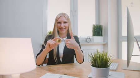 Smiling cheerful blond woman in office suit, sitting at workplace with computer and showing bitcoin in hand doing thumbs up gesture and looking at cameraの写真素材
