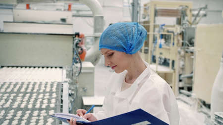 Controller checking conveyor with candies. Young woman in uniform holding folder and inspecting conveyor belt with fresh candies in confectionery factory.の写真素材