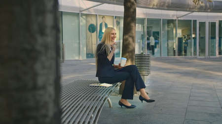 Elegant modern woman sitting on bench and waving with hand to friend while speaking on smartphone outdoorsの写真素材