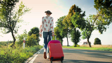Cheerful woman with suitcase walking on road. From below shot of pretty young female smiling and pulling red suitcase while walking on asphalt road during trip through countryside.の写真素材