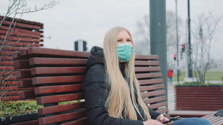 Blonde girl in a medical mask sitting on the bench and using smartphone. She probably reading outbreaking news about COVID-19. Coronavirus pandemic concept.の写真素材