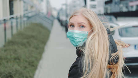 Young blonde woman in a medical mask escaping from somebody on the street. She is scared and concerned. Coronavirus pandemic concept.の写真素材
