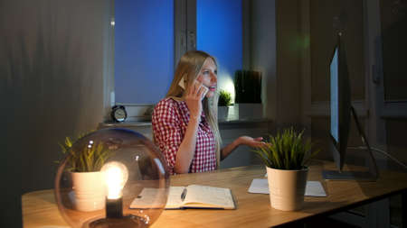 Cheerful amazed blond woman in checkered shirt sitting at night at computer on wooden desk and talking on mobile phone. Happy female at computer talking on smartphone.の写真素材
