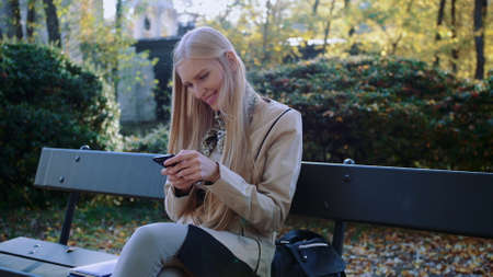 Young blonde girl uses a cell phone on a bench in the Park outdoors.の写真素材