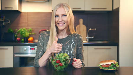 Young lady preferring salad to hamburger. Attractive young woman choosing to eat healthy salad for lunch while sitting at table in stylish kitchen and doing thumbs up gesture and looking at camera.の写真素材