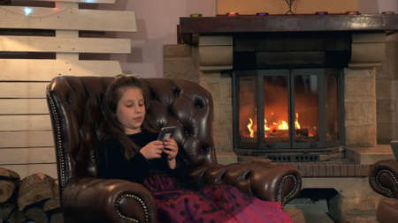 Small girl in nice dress chatting with friends by smartphone in the evening at home. In the background there is a fireplace.の写真素材