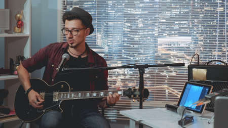 Talented guitarist in hat and trendy casual clothes playing guitar and singing in home recording studio. There are skyscrapers in the background.の写真素材