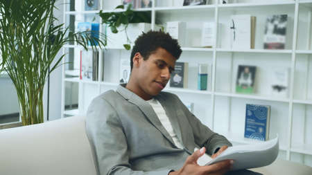 Adult African-American man in suit sitting on sofa in office and looking through paper documents.の写真素材