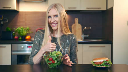 Young lady preferring salad to hamburger. Attractive young woman choosing to eat healthy salad for lunch while sitting at table in stylish kitchen and doing thumbs up gesture and looking at camera.の写真素材