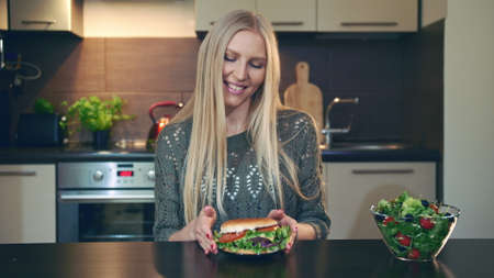 Young lady preferring hamburger to salad. Attractive young woman choosing to eat healthy hamburger for breakfast while sitting at table in stylish kitchen.の写真素材