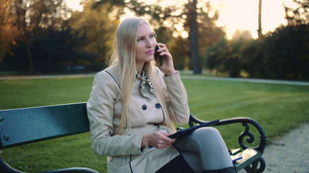 Pretty girl in stylish jacket with tablet computer and cellphone sitting on park bench.の写真素材