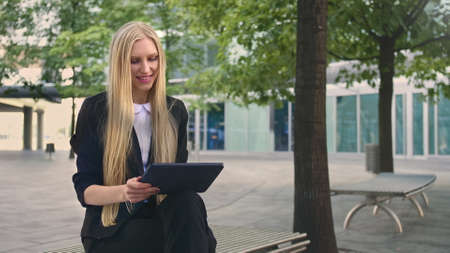 Formal business woman sitting on bench in office patio and browsing tablet in leisure.の写真素材