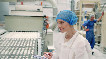Candy factory. Controller checking conveyor with candies. Young woman in uniform holding folder and inspecting conveyor belt with fresh candies in confectionery factory.の写真素材