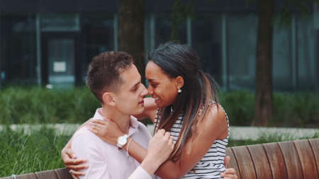 Zoom shot of young mixed-race couple stroking each other sitting on bench outdoors. They having date in a beautiful city park.の写真素材